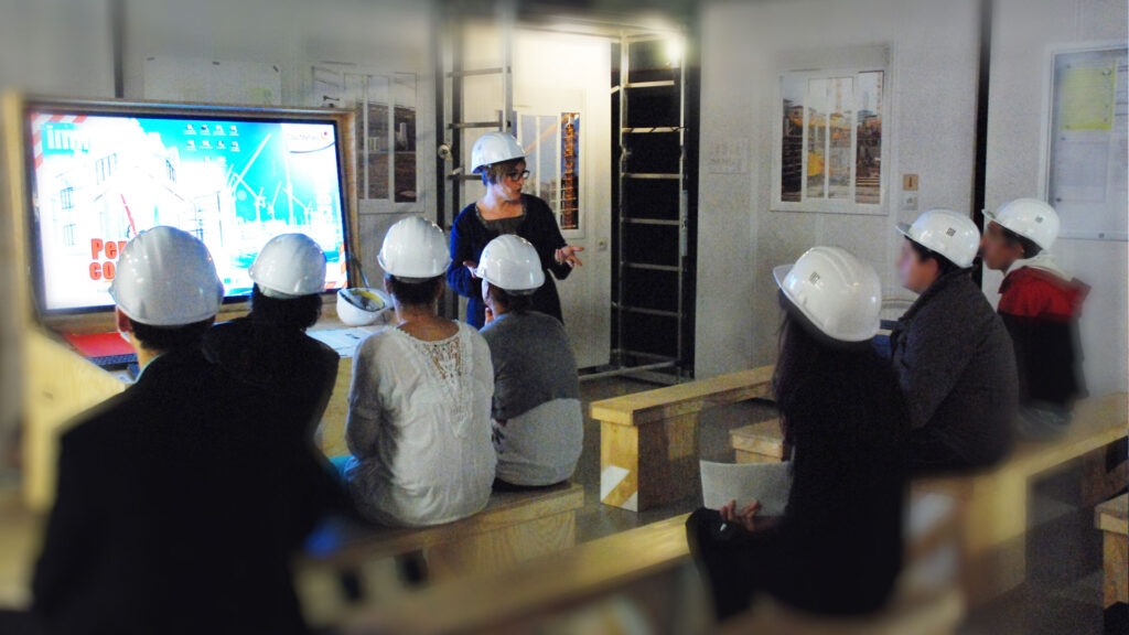Animation pédagogique dans l’exposition « Permis de construire » où un groupe de jeunes en casque de chantier découvre les métiers du bâtiment.