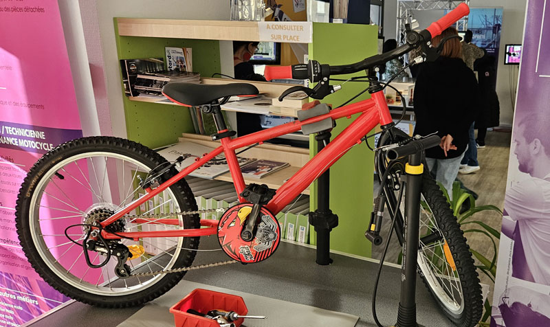 Un vélo est installé sur un pied de réparation avec des outils, dans un espace d’exposition consacré aux métiers de la maintenance et de la mobilité.